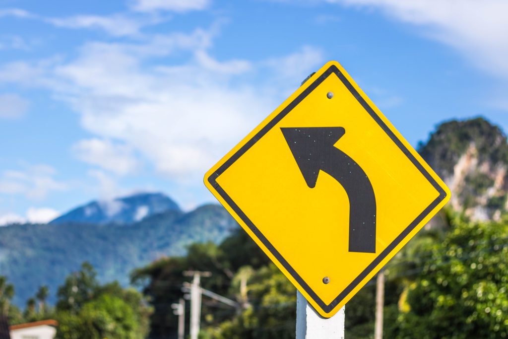 Bright yellow left turn traffic sign with mountain landscape in the background outdoors.